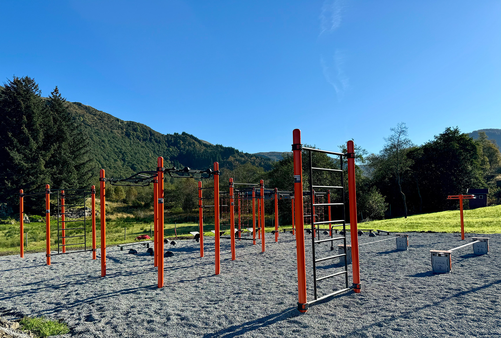 Orange klätterställning och träningsställning på grusplan med skog bakom.